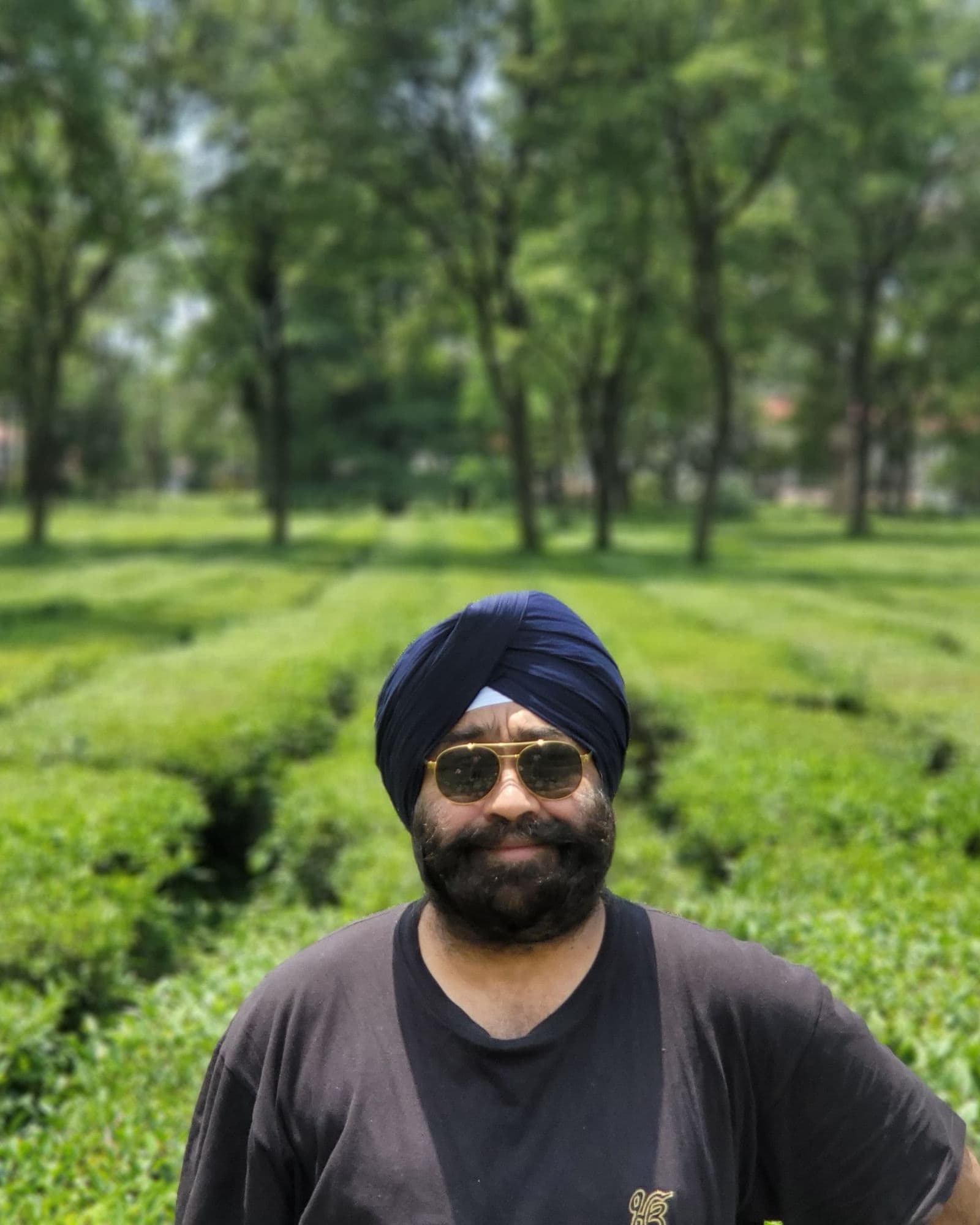 Bupinder Pal Singh Jaggi in a navy turban and sunglasses, smiling, standing between rows of tea bushes