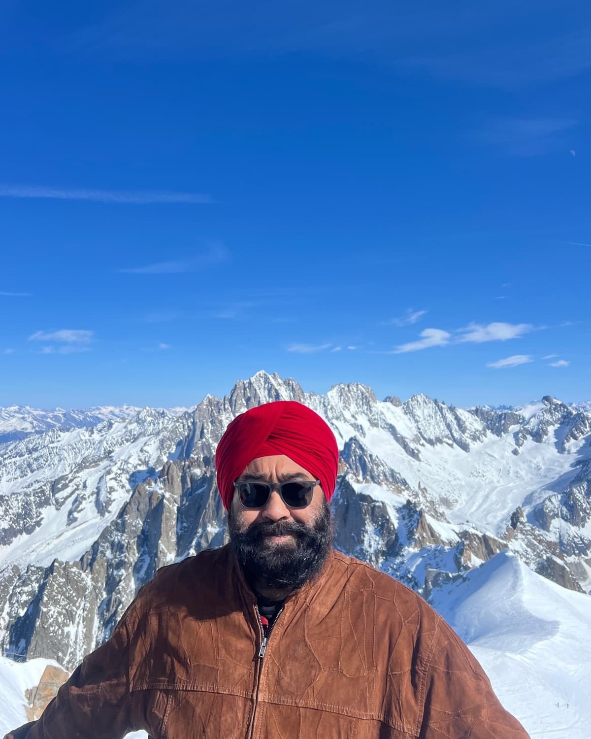 Bupinder Pal Singh Jaggi, founder of Palsee Kashmir, smiling in a red turban and suede jacket with snow-covered mountain peaks behind him
