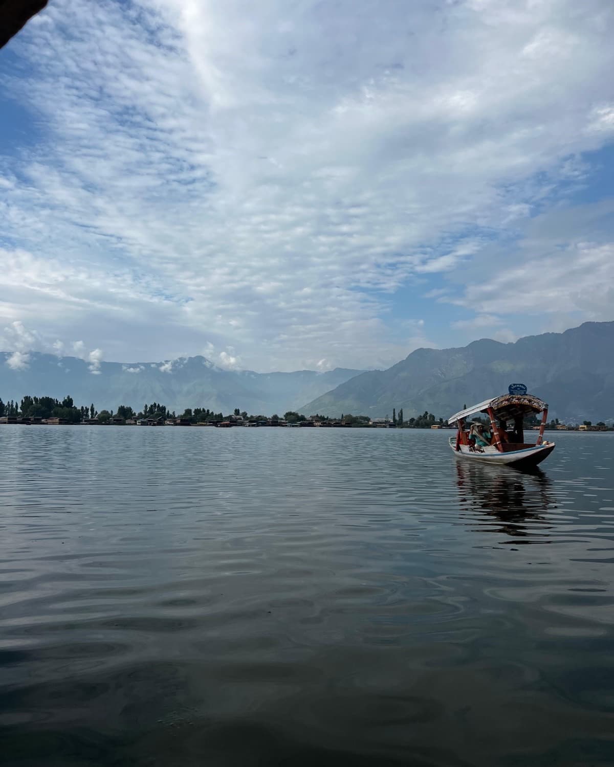 A shikara carrying passengers across Dal Lake, with houseboats along the shore and mountains behind