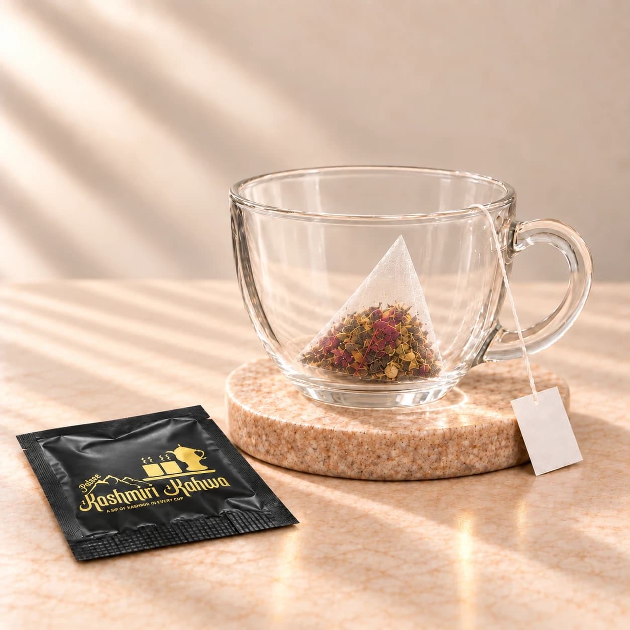 An empty glass cup on a stone coaster with a pyramid tea bag inside, an opened black Kashmiri Kahwa sachet beside it